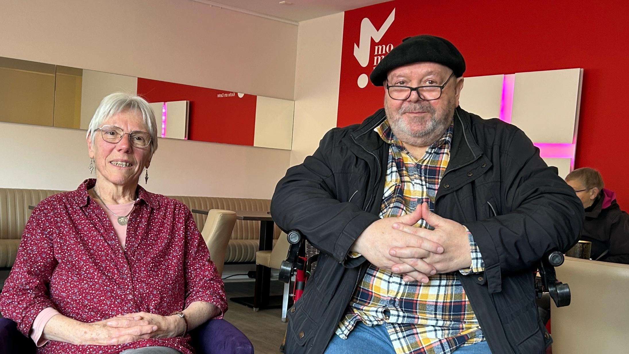 Eine Frau mit Brille sitzt neben einem Mann, der ebenfalls eine Brille und eine Mütze trägt. Beide schauen in die Kamera