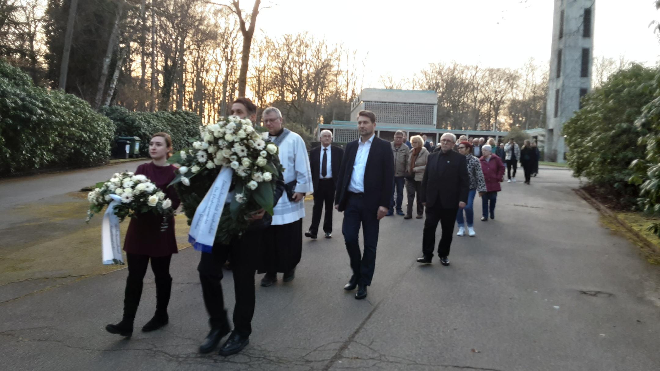 Eine Gruppe von Menschen geht in einem Trauerzug über einen breiten Friedhofsweg. Vorne tragen zwei Personen große weiße Kränze mit Trauerschleifen, begleitet von einer Person in liturgischer Kleidung. Dahinter folgen weitere Teilnehmende in respektvollem Abstand. Rechts im Hintergrund steht ein hoher Glockenturm, links und rechts säumen dichte Sträucher den Weg. Die Abendsonne scheint durch die kahlen Bäume und taucht die Szene in warmes Licht.