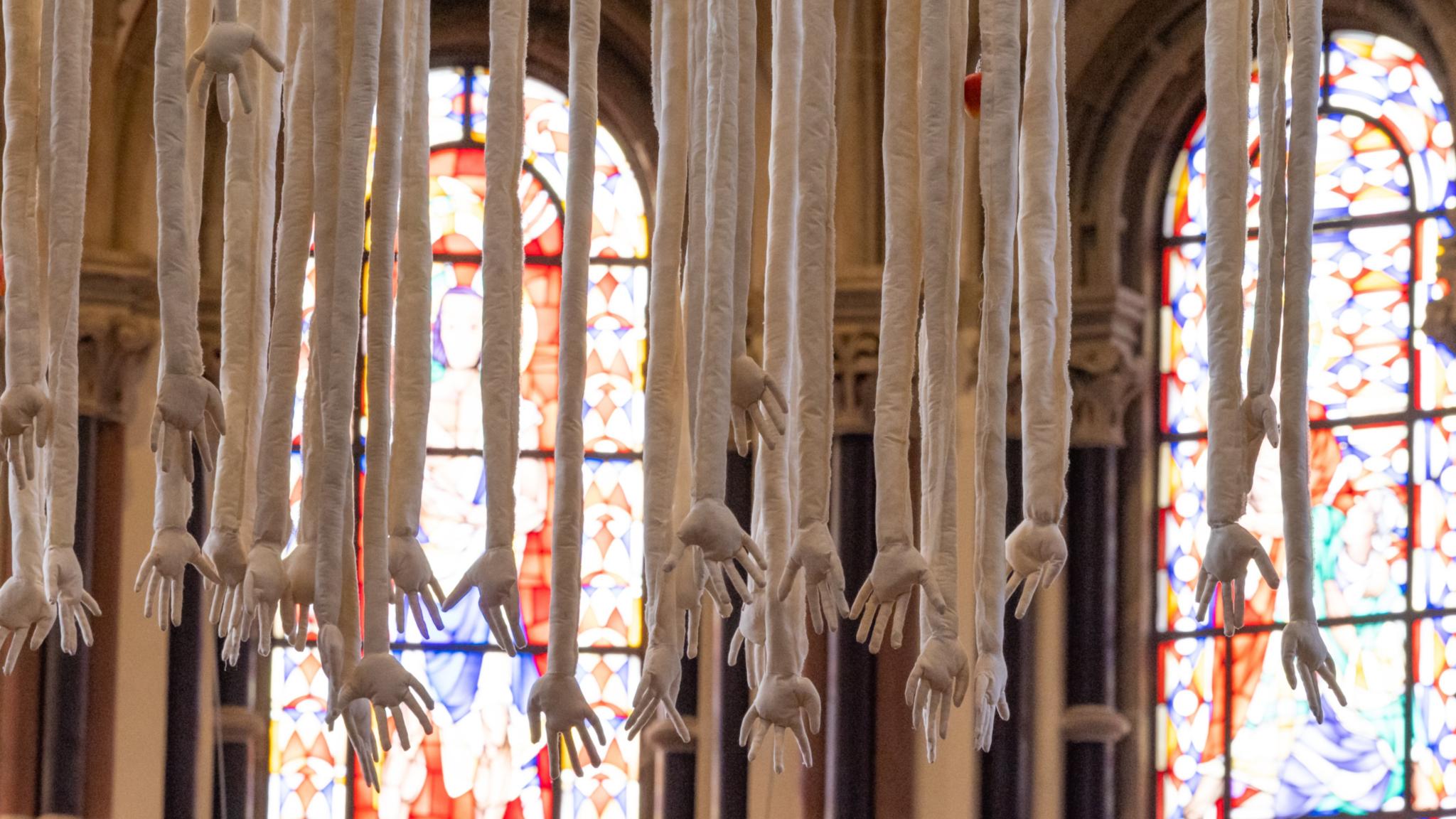 Herabhängende Hände und Arme aus weißem Leinen vor Kirchenfenstern.