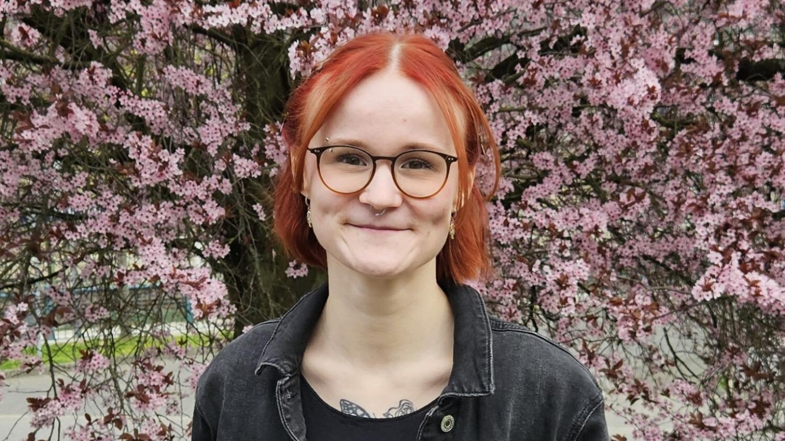 Eine junge Frau mit roten Haare und einer Brille schaut lächelnd in die Kamera.