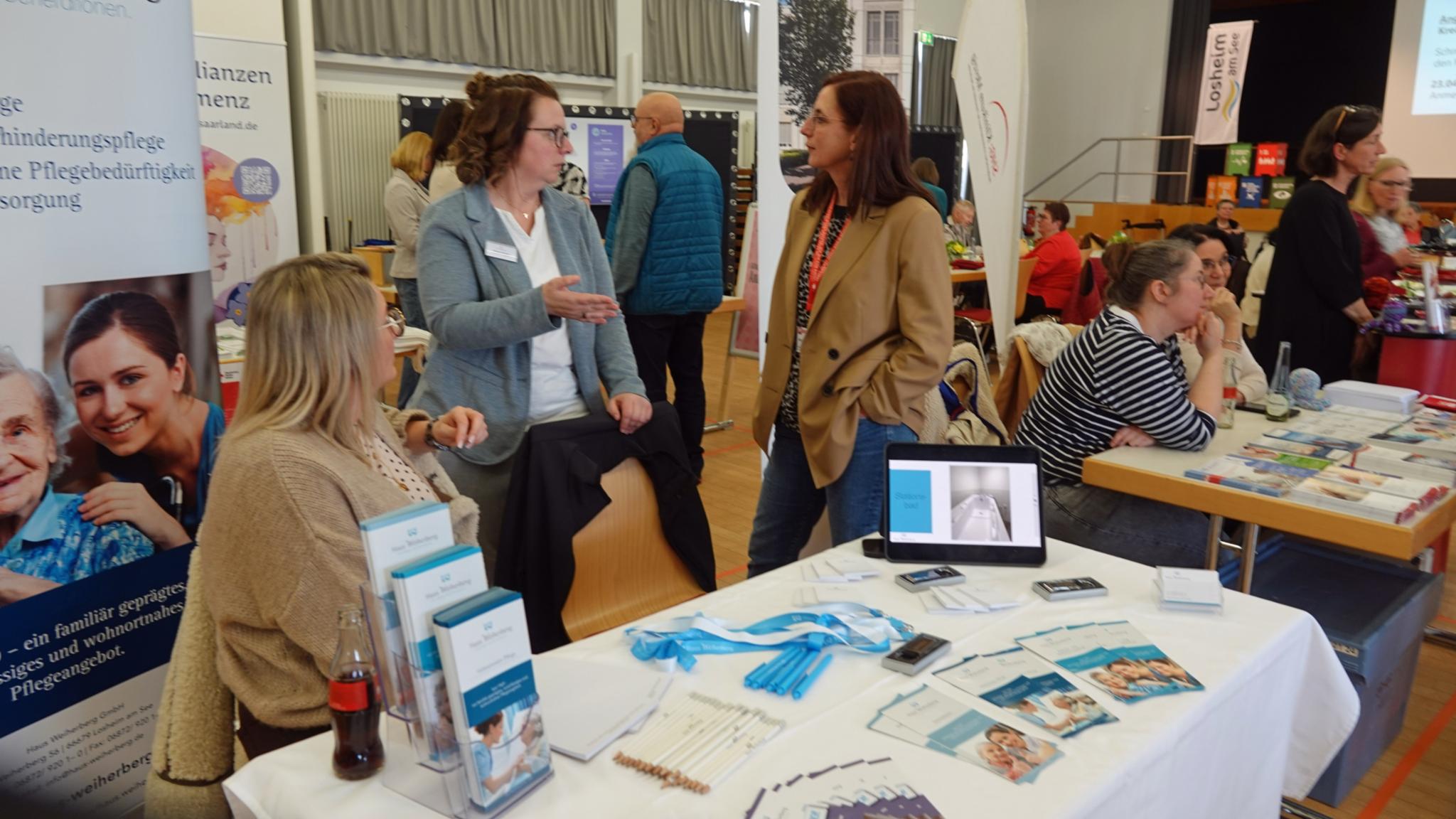In einem großen Saal mit mehreren Informationsständen sitzen und stehen verschiedene Ausstellerinnen und Besucher an Tischen. Im Vordergrund befindet sich ein Stand mit Broschüren, Flyern, Stiften, einem Tablet mit geöffneter Präsentation sowie medizinischem Informationsmaterial. Hinter dem Tisch sprechen mehrere Personen miteinander. Weitere Stände mit Plakaten, Roll-ups und Infomaterial sind im Hintergrund zu sehen. Der Raum ist hell, mit großen Fenstern, Bühnenbereich und zahlreichen Besucherinnen und Besuchern, die sich austauschen und informieren.
