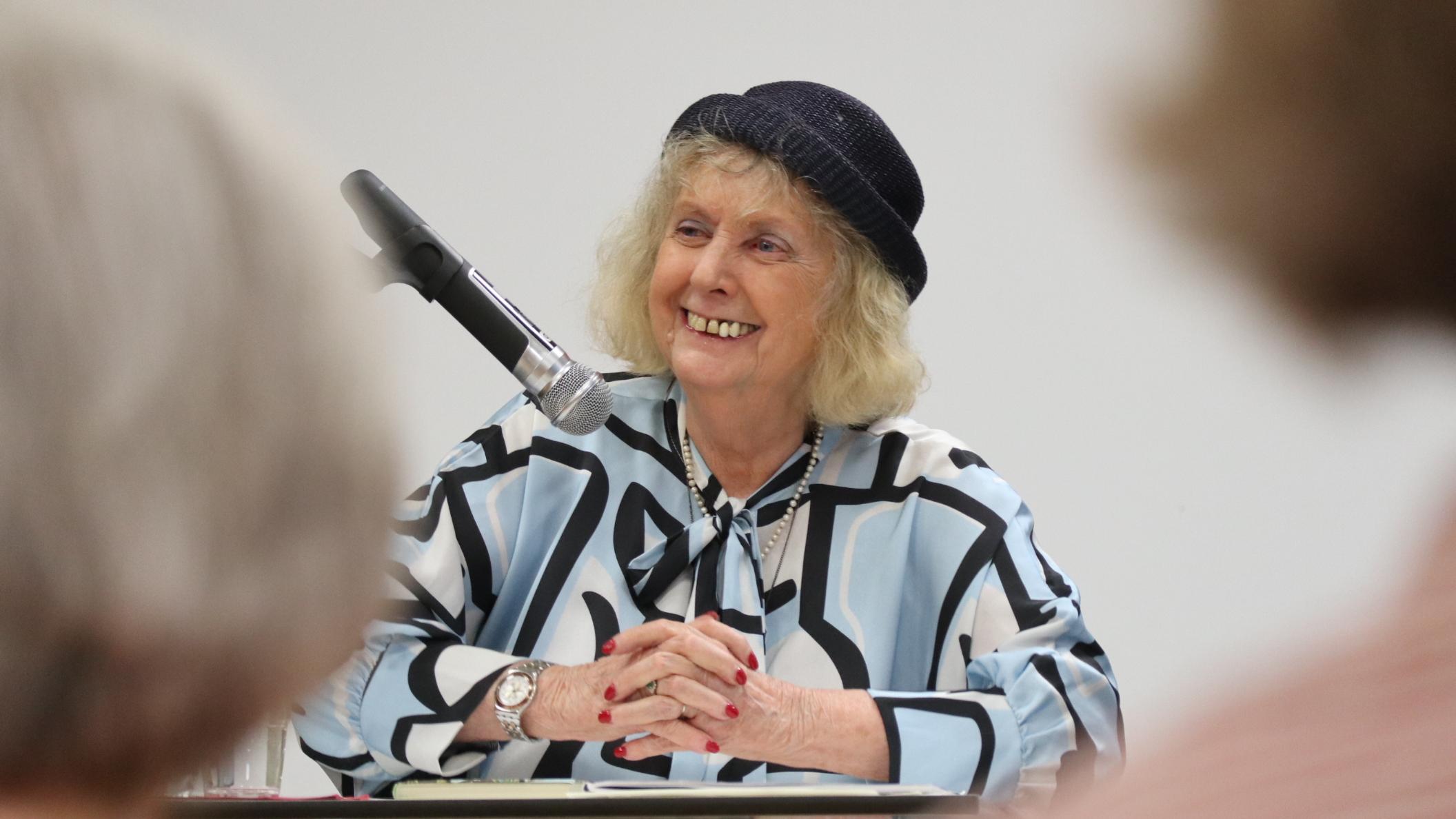 Eine ältere Dame mit Hut sitzt an einem Tisch vor Publikum.